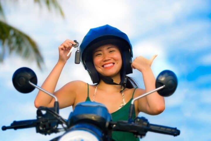 Smiling woman holding scooter key while sitting on a motorcycle with palm trees in the background in Legian Bali &ndash; GoTravela Indonesia