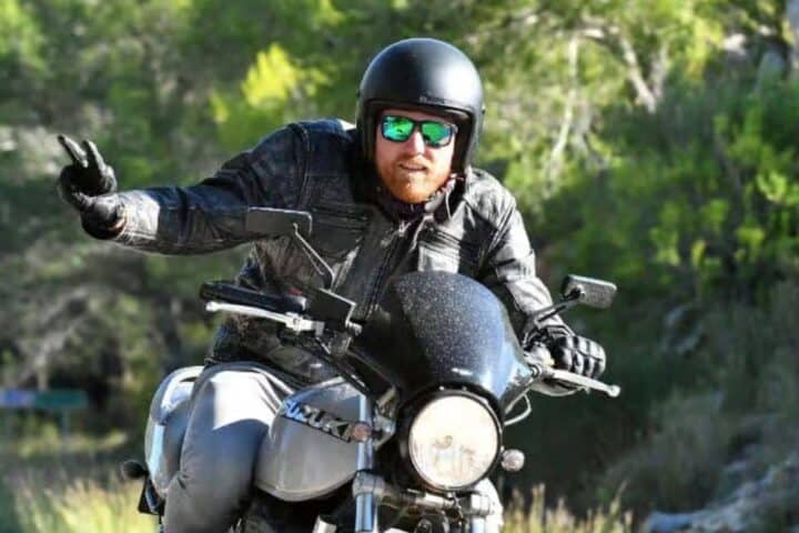 Man riding a black motorcycle wearing helmet and sunglasses on a road surrounded by greenery in Jimbaran Bali &ndash; GoTravela Indonesia